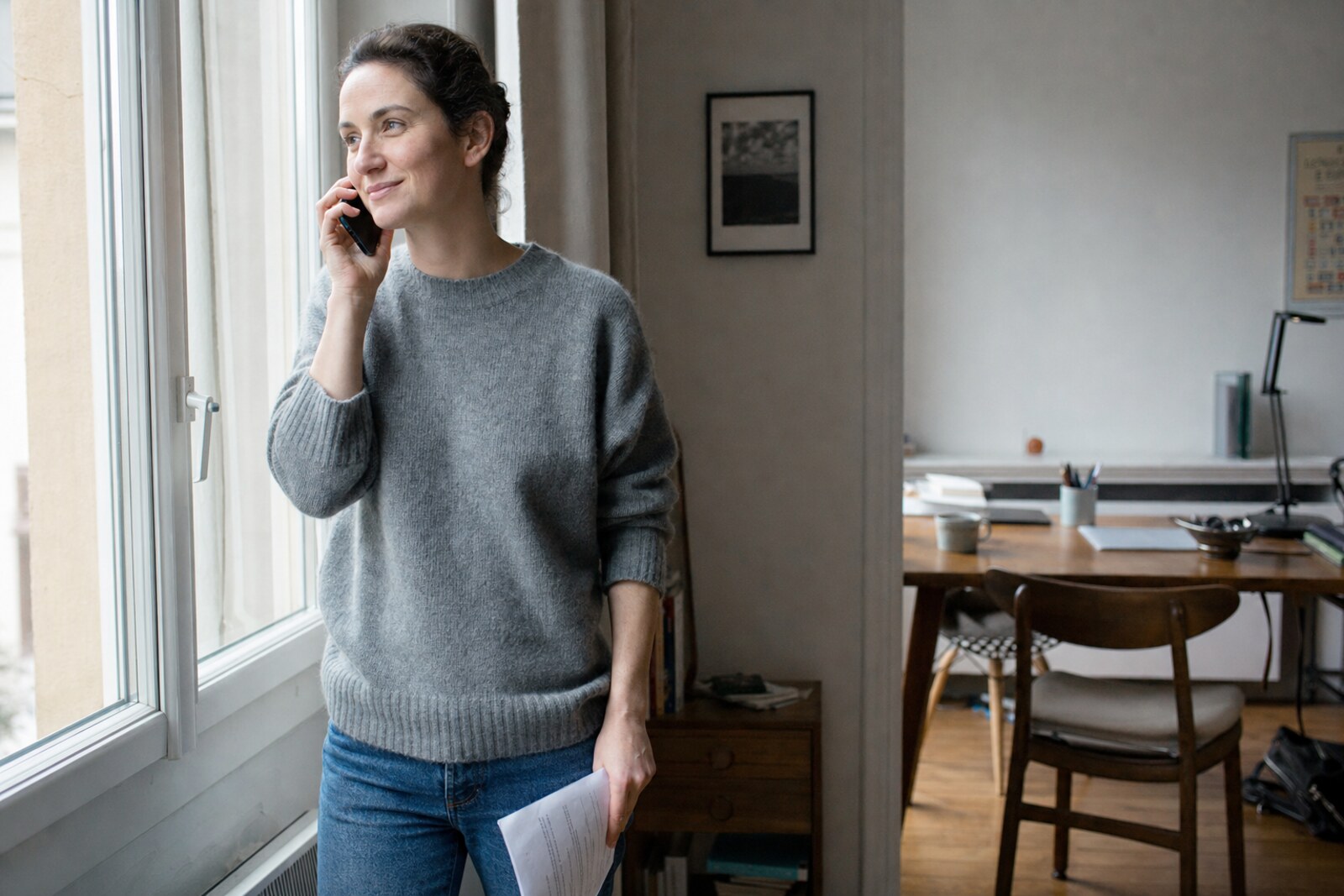 A woman at a bright window on a phone call, holding a printed loan statement